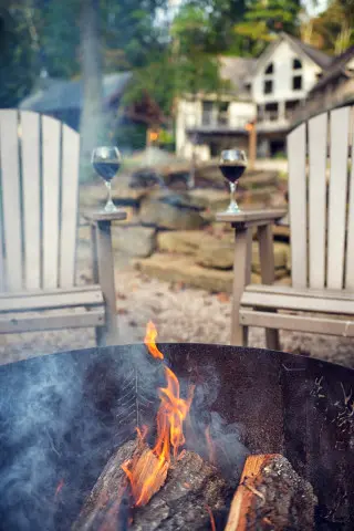 Firepit with logs burning, seats with wine glasses rest on the Adirondack seating, cabin in background 