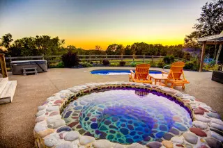 Private stone cold plunge, pool and hot tub in background, sun setting in the sky. 