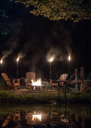 Night time image, with fire pit, tiki torches and Adirondack chairs. Lights reflecting in the water.