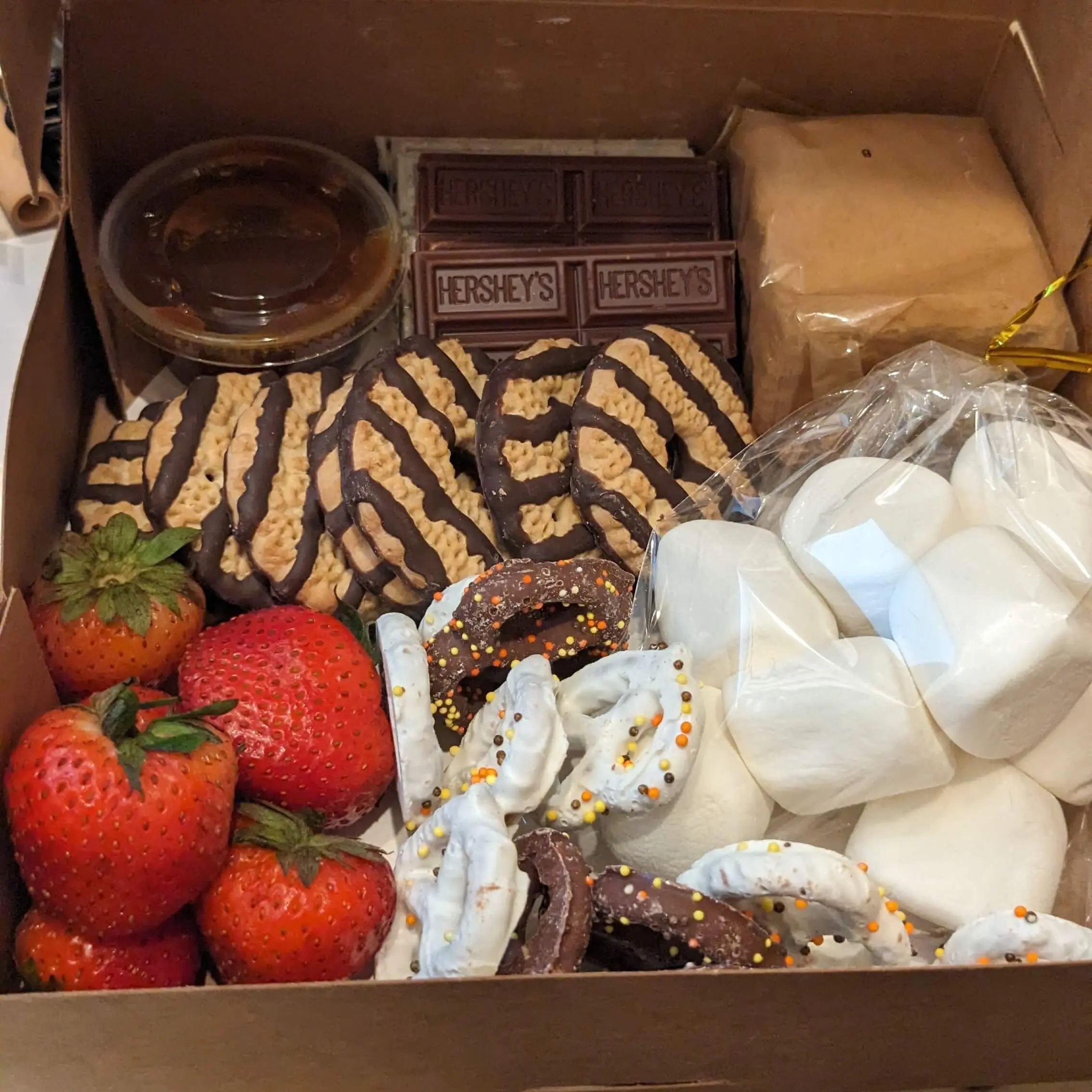 Box of smores supplies, marshmallows, chocolate, strawberries, caramel sauce, graham crackers, chocolate covered pretzels, cookies. 