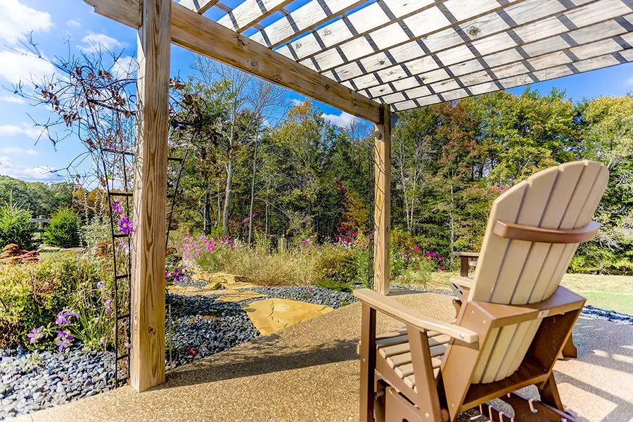 A chair beneath a wooden pergola overlooking a landscaped garden and wooded hillside.