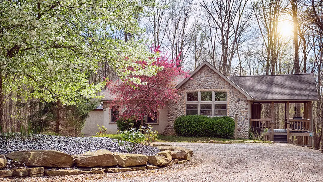 Rustic stone cabin surrounded by trees with a gravel driveway and covered porch.