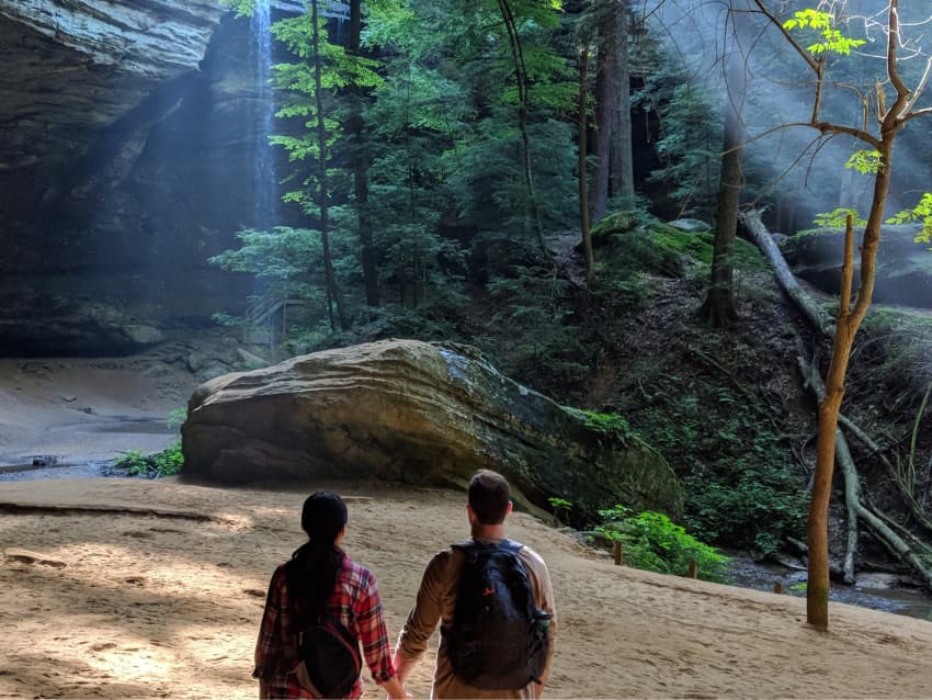 Couple standing hand-in-hand, looking at the stunning Ash Cave at Hocking Hills in the background