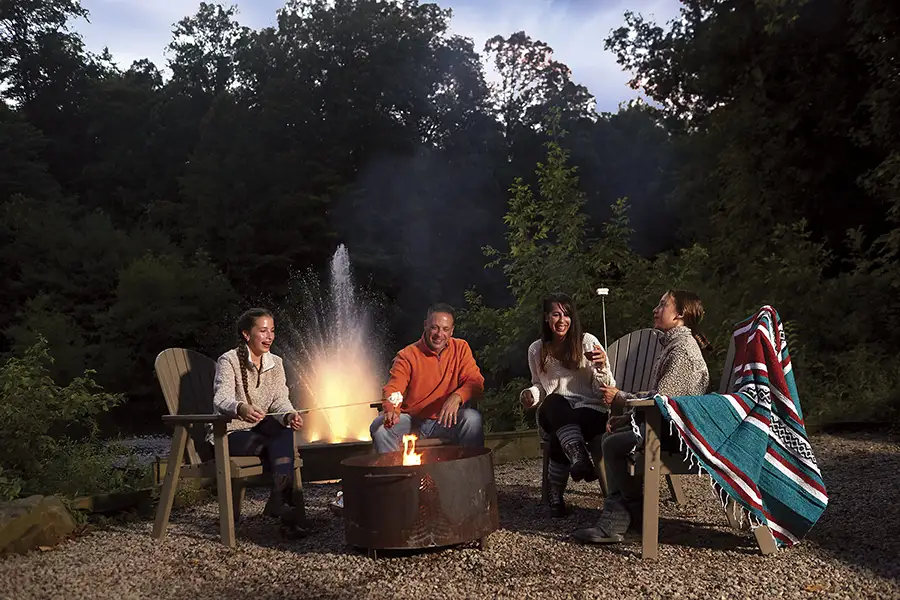 Family and friends roasting marshmallows around a backyard fire pit surrounded by trees.
