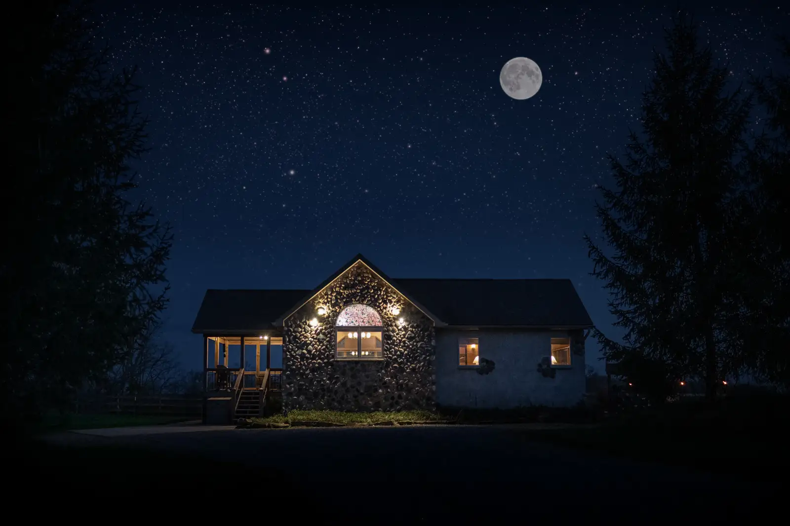Cabin illuminated at night beneath a star-filled sky and bright moon, framed by tall trees.