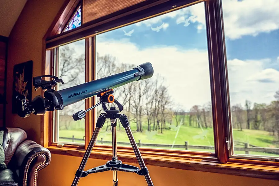 Telescope Overlooking a Country Landscape Telescope on a tripod positioned beside large windows overlooking a green rural landscape and blue sky.