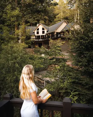 Woman with a book stands at a dock, looking at the large cabin in the background