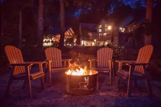 A fire burns in a fire pit, 4 Adirondack chairs surround. Cabin glows with warm light in the background 