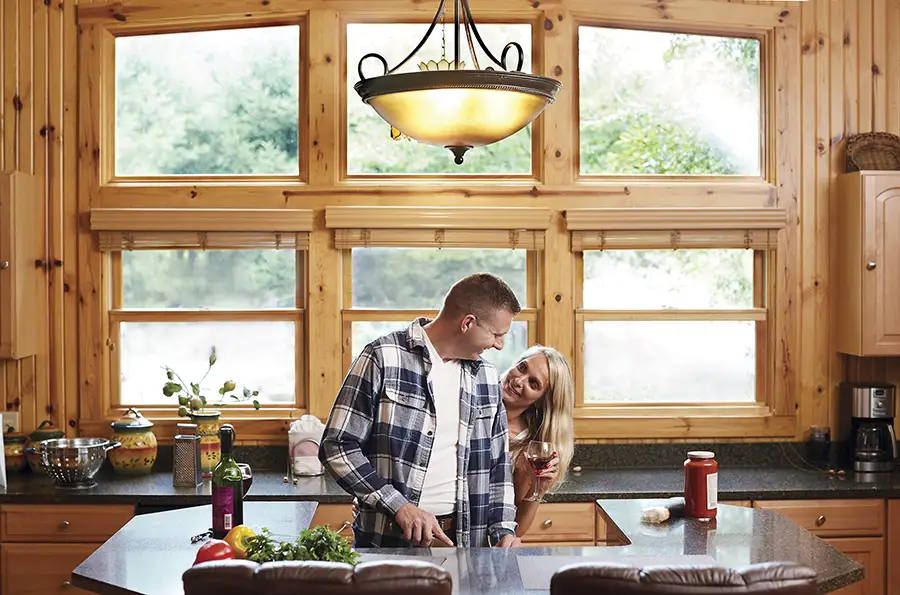 A smiling couple prepares fresh vegetables and shares a glass of wine in a warm, wood-paneled kitchen.