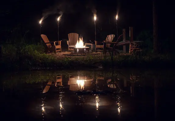 Night time image, with fire pit, tiki torches and Adirondack chairs. Lights reflecting in the water.