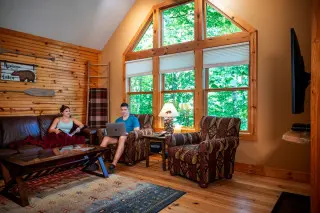 Woman sits on a couch with a throw over her. Man sits in an armchair on a laptop. Large windows in the background show scenic nature.