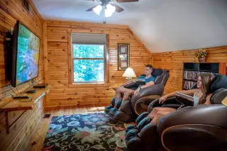 Man and woman sit in massage chairs, watching the mounted tv. 