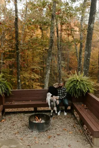 Couple sitting close on benches surrounding the fire pit, fall colors on trees in the background