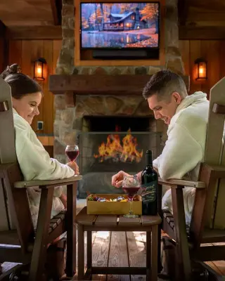 a man and woman sit in robes in front of a fireplace. They have glasses of wine and are eating charcuterie