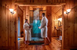 A man and woman stand in robes outside of a glass shower, stained glass in the background