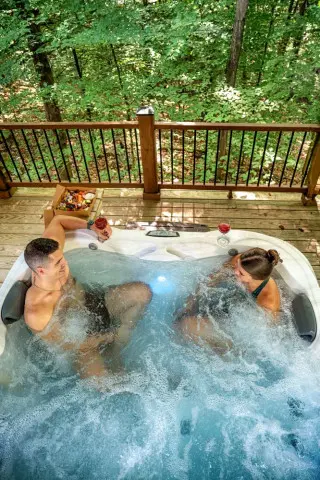 A man and woman sit in a bubbling hot tub. A box of charcuterie on a chair nearby, forest views in background