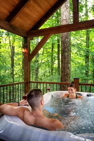 A woman and man sit in a hot tub, with forest views surrounding