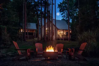 Night scene, cabin with warm lights glowing in the background, fire in the firepit with seating surrounding
